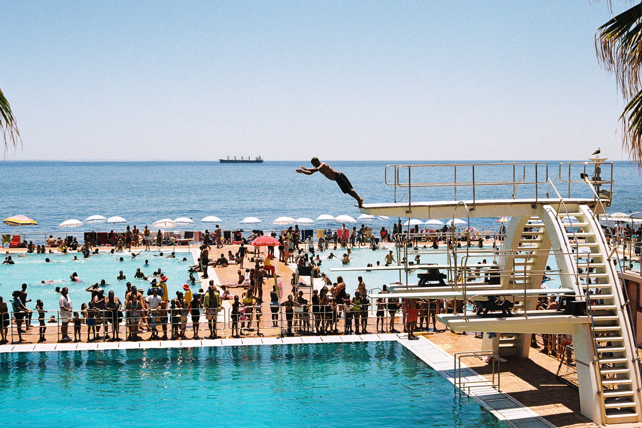 Person diving into a pool with a beach and ocean view, slim aarons style photograph